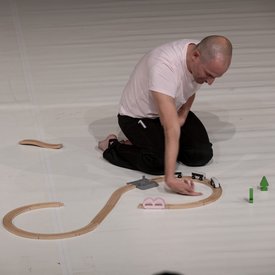 Michael Turinsky auf der Bühne in einer Tanzpose auf den Knien. Er trägt ein helles Shirt und eine schwarze Hose. Vor ihm eine halb zusammengebaute Spielzeug-Holzeisenbahn.