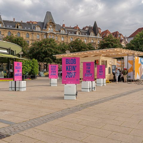 Foto der Ausstellung »Schlamassel Tov« auf dem Marienplatz vor der Sukka: Mehrere pinke Plakate mit blauer Aufschrift stehen vor einem Bau aus Holz und Stoff – der Laubhütte bzw. Sukka von Sukkat Salaam. Im Vordergrund ist ein Plakat mit der Aufschrift »Bloß kein‘ Zoff« zu sehen.