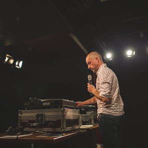 Pascal Claude steht an seinem DJ-Pult. In der einen Hand hält er ein Mikrofon. Bekleidet ist er mit einer Jeans und einem karierten Hemd.