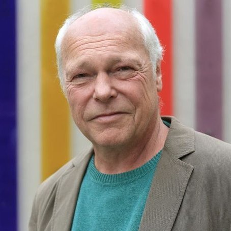 Portraiaufnahme von Professor Schumacher. Professor Schumacher hat kurze, fast weiße Haare und trägt auf dem Bild ein türkisfarbenes Shirt und ein beiges Jackett.