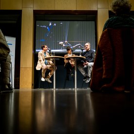 Blick auf das Podium mit Moderator und den beiden Gäst*innen. Der Fotograf hat das Bild aus den zuschauerreihen heraus aufgenommen, so dass am rechten und linken Bildrand Zuschauer zu erkennen sind.