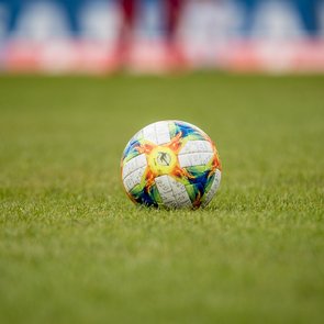 Ein bunter Fußball liegt auf einer grünen Rasenfläche. Der Ball zeigt ein auffälliges Muster mit roten, gelben, blauen und grünen Farben. Der Hintergrund ist unscharf und zeigt Teile eines Fußballfelds oder Stadions.“