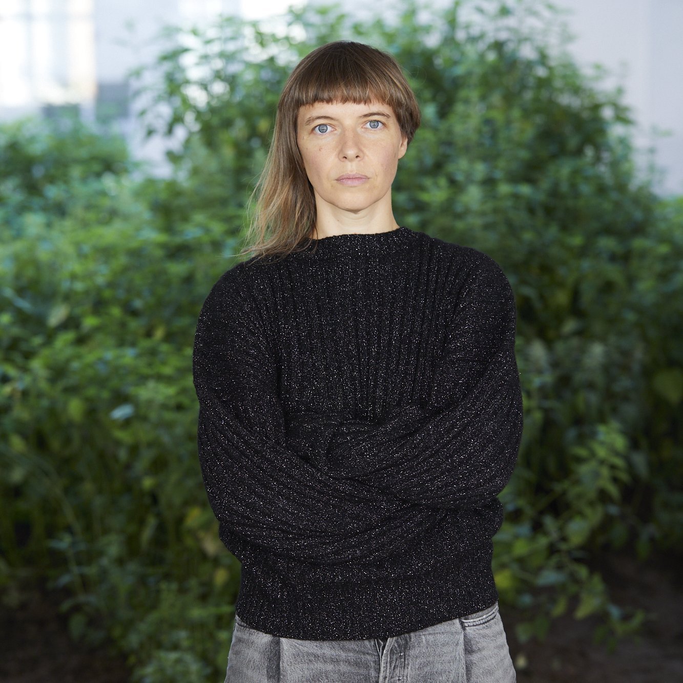 Portraitaufnahme von Lina Lapelytè vor einem Busch. Lina Lapelytè hat lange Haare und einen geraden Pony. Bekleidet ist sie mit einem schwarzen Pulli und einer grauen Jeans.