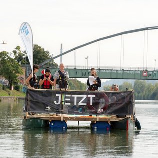 Das Festival-Floß unterwegs auf einem Gewässer in der Region. An Bord sind verschiedene Personen. Ein Banner an der Reling des Floßes zeigt das Logo von JETZT!.