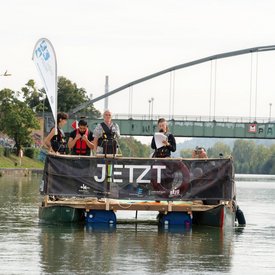 Das Festival-Floß unterwegs auf einem Gewässer in der Region. An Bord sind verschiedene Personen. Ein Banner an der Reling des Floßes zeigt das Logo von JETZT!.