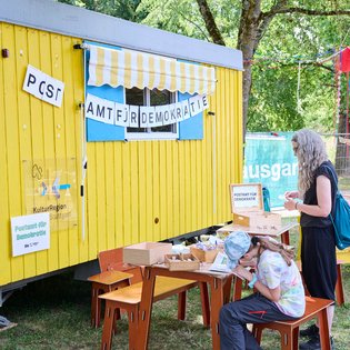 vor einem gelben Bauwagen mit der Aufschrift "Postamt für Demokratie" sitzen Menschen auf Holzbänken, Erwachsene und Kinder und basteln am Tisch