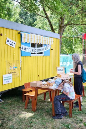 vor einem gelben Bauwagen mit der Aufschrift "Postamt für Demokratie" sitzen Menschen auf Holzbänken, Erwachsene und Kinder und basteln am Tisch