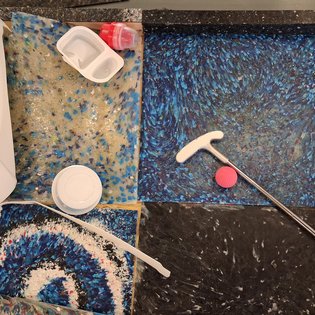 Mehrfarbige Bodenfliesen mit Marmorierung, darauf liegen ein Golfschläger, ein roter Golfball, ein weißer Becher, ein weißer Behälter mit rotem Deckel und ein weißes Tuch