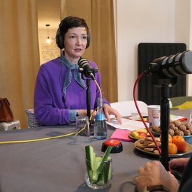 Kimsy von Reischach, Person mit lila Pullover und blauem Halstuch sitzt an Tisch mit Mikrofon, Kopfhörern, Wasserflasche, Obst und Keksen.