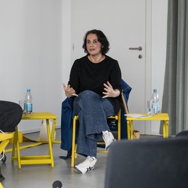 Johara Berriane sitzt auf einem gelben Stuhl und scheint mit jemandem zu sprechen. Mit den Händen gestikuliert sie. Sie hat halblange, braune Haare. Neben ihr stehen zwei kleine gelbe Tischchen.