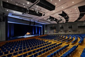 Innenansicht auf den bestuhlten Kombisaal. Die Wände sind in hellen Tönen gehalten, der Saal verfügt über einen Parkettboden. Hinten im Saal ist eine Bühne zu sehen