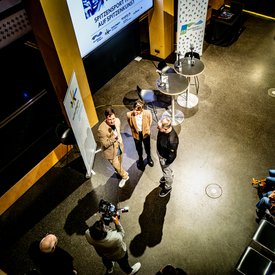Moderator Ingmar Volkmann, Rocío Alemán und Alexander Zorniger stehen vor der eigentlichen Podcast-Aufnahme beieinander und sprechen miteinander. Sie werden dabei von einem Kameramann gefilmt. Fotografiert wurde von der Galerie im ersten Stock des Gebäudes aus.