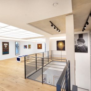 Innenraum einer Galerie im Fritz Genkinger Kunsthaus mit hellen Wänden, Holzfußboden und moderner Beleuchtung. An den Wänden hängen farbenfrohe Gemälde, im Vordergrund ein schwarzes Metallgeländer um das offene Treppenhaus. Rechts ein großes Porträtfoto von Fritz Genkinger sowie eine Wand mit biografischem Text über den Künstler.