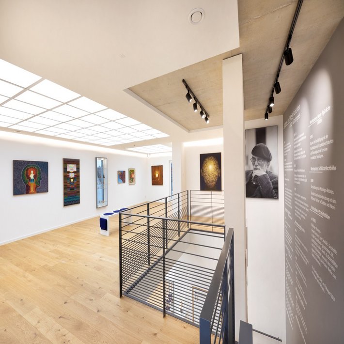 Innenraum einer Galerie im Fritz Genkinger Kunsthaus mit hellen Wänden, Holzfußboden und moderner Beleuchtung. An den Wänden hängen farbenfrohe Gemälde, im Vordergrund ein schwarzes Metallgeländer um das offene Treppenhaus. Rechts ein großes Porträtfoto von Fritz Genkinger sowie eine Wand mit biografischem Text über den Künstler.