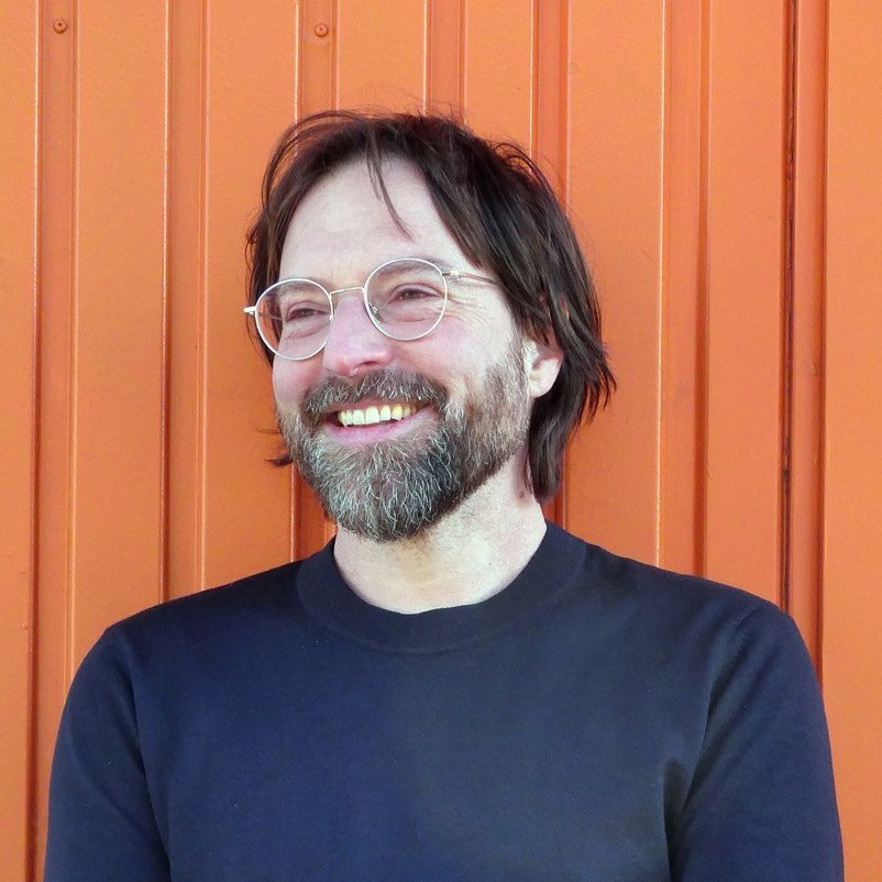 Mann mit Brille und Bart lächelt vor orangefarbener Wand.