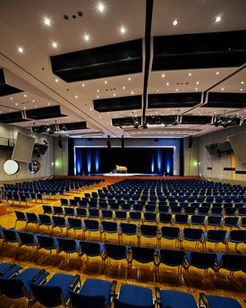 Innenansicht auf den bestuhlten Kombisaal. Die Wände sind in hellen Tönen gehalten, der Saal verfügt über einen Parkettboden. Hinten im Saal ist eine Bühne zu sehen