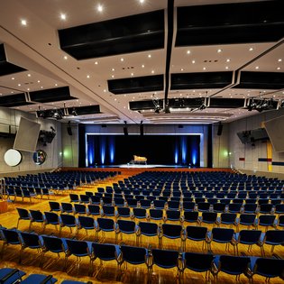 Innenansicht auf den bestuhlten Kombisaal. Die Wände sind in hellen Tönen gehalten, der Saal verfügt über einen Parkettboden. Hinten im Saal ist eine Bühne zu sehen