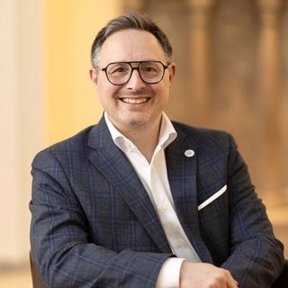 Portraitbild von Michael Rubinstein. Er lächelt in die Kamera, trägt einen dunklen Anzug, ein weißes Hemd und eine Brille. Der Hintergrund ist nur verschwommen dargestellt.