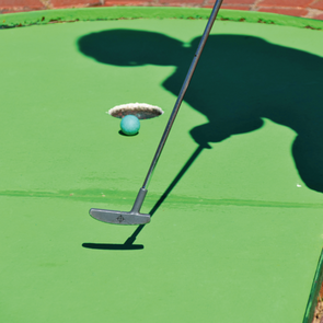 Minigolf-Putter trifft blauen Ball auf grünem Spielfeld mit Schatten einer Person im Hintergrund, dargestellt in einem kreisförmigen Ausschnitt, der wie ein Loch wirkt.