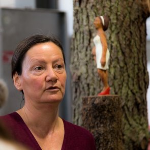 Künstlerin Birgit Refeldt vor einer Skulptur an einem Baumstamm. Frau Refeldt trägt die Haare nach hinten gekämmt, ist mit einem weinroten Oberteil bekleidet und schein zu jemandem zu sprechen.