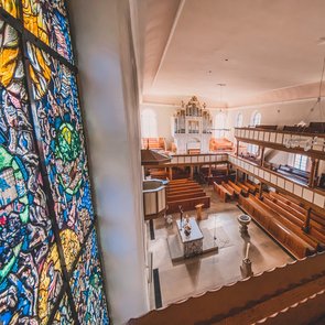 Blick in die Kirche. Zu sehen sind Buntglasfenster, Holzbänke und der Altarraum