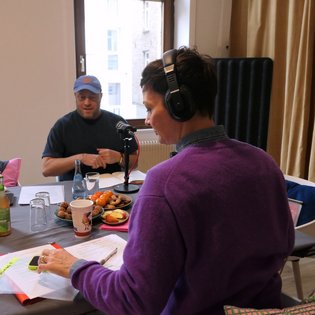 Zwei Personen, Kimsy von Reischach und Jacob Bilabel, sitzen an einem Tisch mit Mikrofonen, Getränken und Snacks, eine Person trägt Kopfhörer und einen lila Pullover.