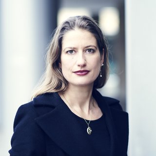 Portraitaufnahme von Tina Hartmann. Tina Hartmann hat langes, blondes Haar, ist mit einem dunkelblauen Shirt und einer ebenso dunkelblauen Jacke bekleidet, die sie mit einer langen Kette mit Anhänger kombiniert.