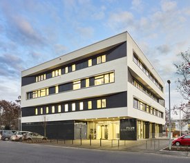 Modernes Bürogebäude mit weißer Fassade, schwarzen Fensterbändern und dem Schriftzug »FILUM« an der Ecke. Mehrere Autos parken entlang der Straße