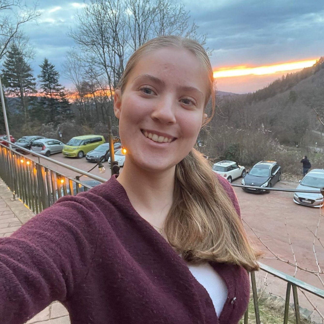 Junge Frau mit langem blonden Haar und bordeauxrotem Pullover macht ein Selfie auf einem Balkon mit Blick auf geparkte Autos und bewaldete Hügel im Sonnenuntergang