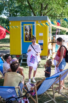 vor einem gelben Bauwagen mit der Aufschrift "Postamt für Demokratie" sitzen Menschen auf Holzbänken, Erwachsene und Kinder und basteln am Tisch
