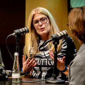 Nina Preuss (Agentur Preuss und Preuss) hat blondes, langes Haar und trägt ein schwarz-weißes Shirt mit Aufdruck 'STUTTGART'. Sie gestikuliert mit beiden Händen vor zwei Mikrofonen.