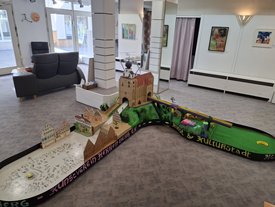 Minigolfbahn mit Modellgebäuden und einer Burg in einem hellen Raum mit Sitzmöbeln und Bildern an den Wänden