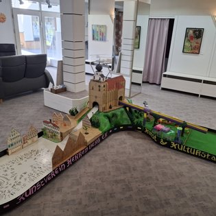 Minigolfbahn mit Modellgebäuden und einer Burg in einem hellen Raum mit Sitzmöbeln und Bildern an den Wänden