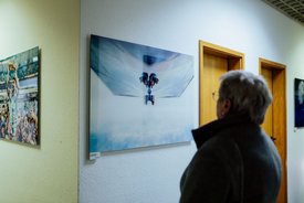 Person mit Brille und grauen Haaren betrachtet ein Foto an einer weißen Wand mit zwei weiteren Fotos in einem Flur mit Holztüren