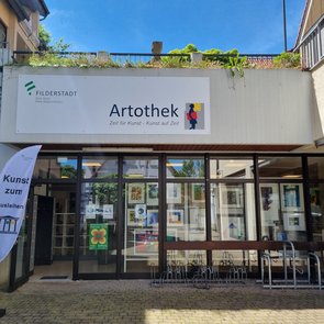 Eingangsbereich der Artothek Filderstadt bei sonnigem Wetter. Über dem Eingang hängt ein großes Schild mit dem Logo der Stadt Filderstadt und dem Schriftzug „Artothek – Zeit für Kunst – Kunst auf Zeit“. Links neben der Tür steht eine weiße Fahne mit der Aufschrift „Kunst zum Ausleihen“. Durch die Glasfront sind zahlreiche gerahmte Kunstwerke im Innenraum sichtbar. Vor dem Eingang befinden sich Fahrradständer.