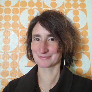 Person mit kurzem, gewelltem Haar vor einer Wand mit orange-weißem geometrischem Muster