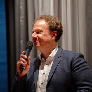 Dr. Matthias Knecht eröffnet den Kurzfilmnachmittag. Er steht auf der Bühne vor der Leinwand und hält ein Mikrofon in der Hand.