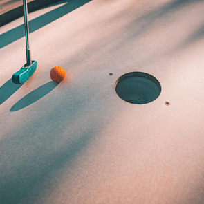 Minigolfball neben Loch auf Minigolfbahn mit Schläger und Schatten