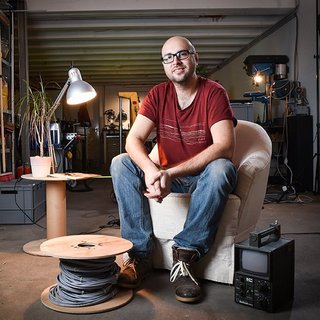 Ein Mann sitzt in einem weißen Sessel in einer Werkstatt. Er trägt ein rotes T-Shirt und Jeans. Neben ihm stehen ein Tisch mit Pflanze und Lampe sowie eine Kabelrolle und ein elektronisches Gerät. Im Hintergrund sind Werkzeuge und Regale zu sehen.
