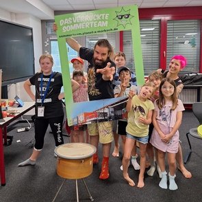Gruppe von Kindern mit einem Erwachsenen, einige halten einen großen grünen Rahmen mit der Aufschrift 'DAS VERRÜCKTE SOMMERTEAM', dargestellt in einem kreisförmigen Ausschnitt, der wie ein Loch wirkt.