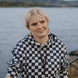 Person mit kurzem, blondem Haar trägt einen schwarz-weißen Kapuzenpullover mit Schachbrettmuster, im Hintergrund Wasser und Felsen.
