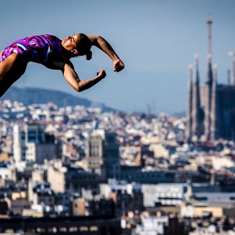 Sportlerin in violettem Badeanzug springt vom Sprungbrett vor einer Stadtlandschaft mit der Sagrada Família im Hintergrund
