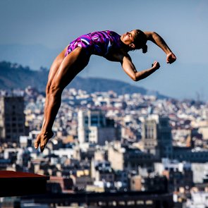 Sportlerin in violettem Badeanzug springt vom Sprungbrett vor einer Stadtlandschaft mit der Sagrada Família im Hintergrund