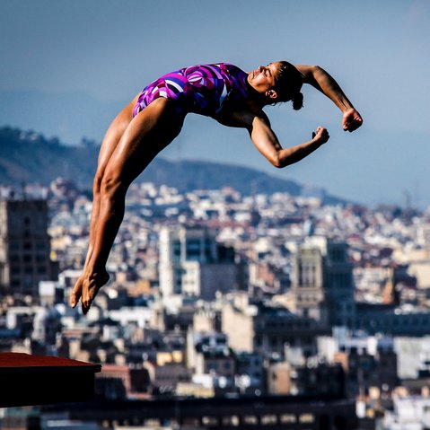 Sportlerin in violettem Badeanzug springt vom Sprungbrett vor einer Stadtlandschaft mit der Sagrada Família im Hintergrund