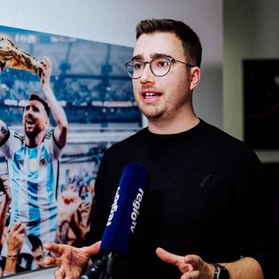 Mann in schwarzem Pullover hält ein blaues Mikrofon mit der Aufschrift 'regio tv' vor einem großen Bild der Fußball-Weltmeisterschaft mit jubelnden Spielern in hellblauen und weißen Trikots.