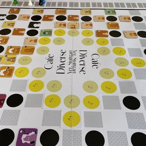 Ein buntes Brettspiel mit dem Titel „Café Diverse“ ist auf einem Tisch aufgebaut. Das Spielfeld zeigt viele farbige Kreise und Quadrate in Gelb-, Grau- und Schwarztönen, auf denen kleine quadratische Plättchen mit Comicfiguren liegen. Um das Spielfeld herum stehen Spielfiguren in verschiedenen Farben (Gelb, Grün, Weiß). Am Rand befindet sich eine Zählleiste mit Zahlen. In der Mitte des Spielfelds steht mehrfach der Schriftzug „Café Diverse – Yes, Sürgent Yes! Stuttgart“.