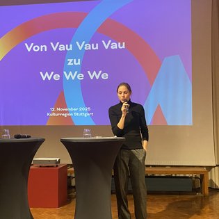 Bettina Pau, Geschäftsführerin der KulturRegion Stuttgart, begrüßt das Publikum. Im Hintergrund eine Präsentation über "Von Vau Vau Vau zu We We We".