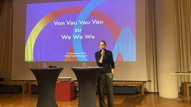 Bettina Pau, Geschäftsführerin der KulturRegion Stuttgart, begrüßt das Publikum. Im Hintergrund eine Präsentation über "Von Vau Vau Vau zu We We We".