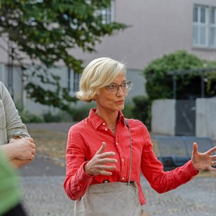 Eine ältere Teilnehmerin in einem roten schicken Hemd spricht auf einem Platz in Zuffenhausen zu den anderen Teilnehmern und gestikuliert mit offenen Armen