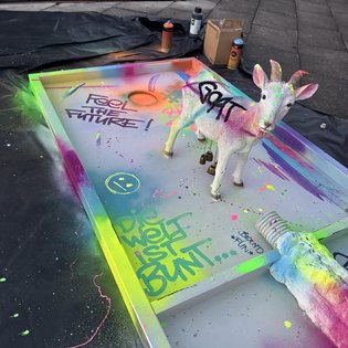 Minigolfbahn mit buntem Graffiti, darauf eine weiße Ziegenskulptur mit Schriftzug und bunte Farbspritzer